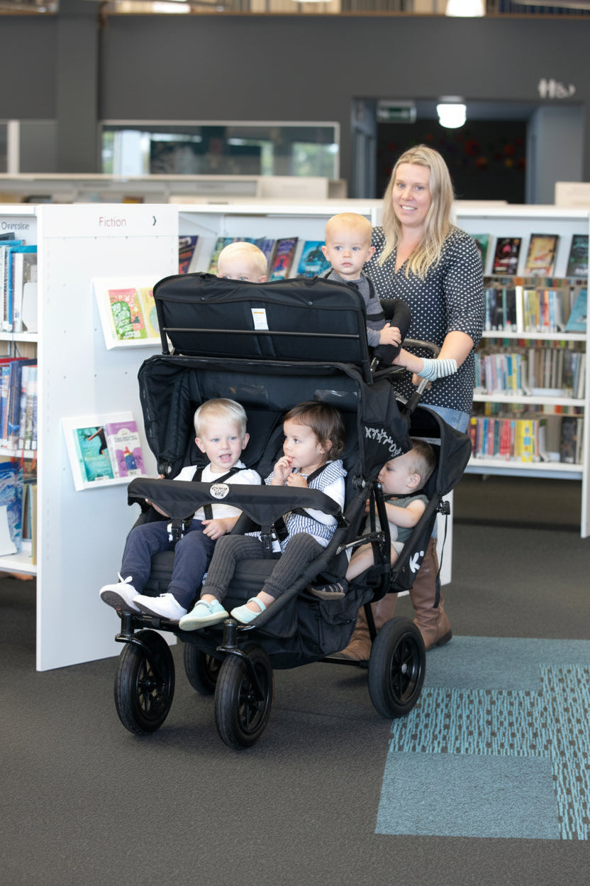 A multi-seat push chair with six children been pushed by a childminder in a library